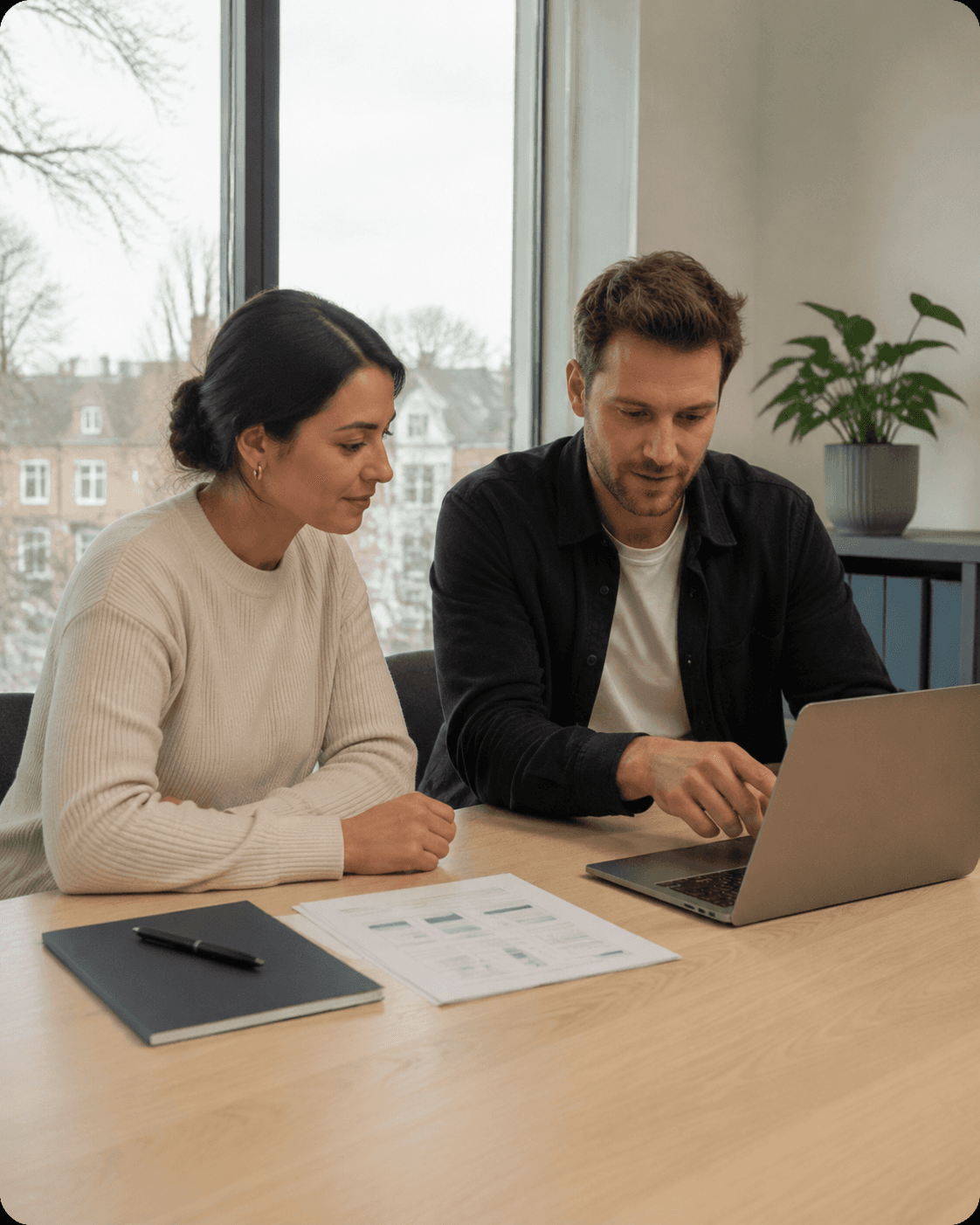Man and woman sitting at a desk, looking at a laptop screen in an office.